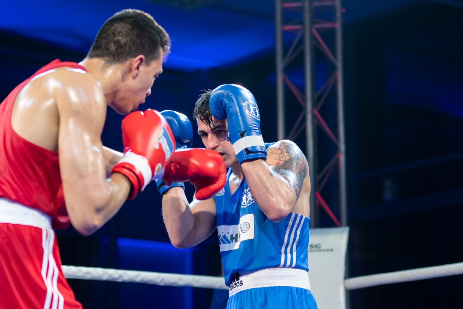 Zwei Boxer in roter und blauer Sportkleidung stehen sich mit erhobenen Handschuhen im Ring gegenüber.