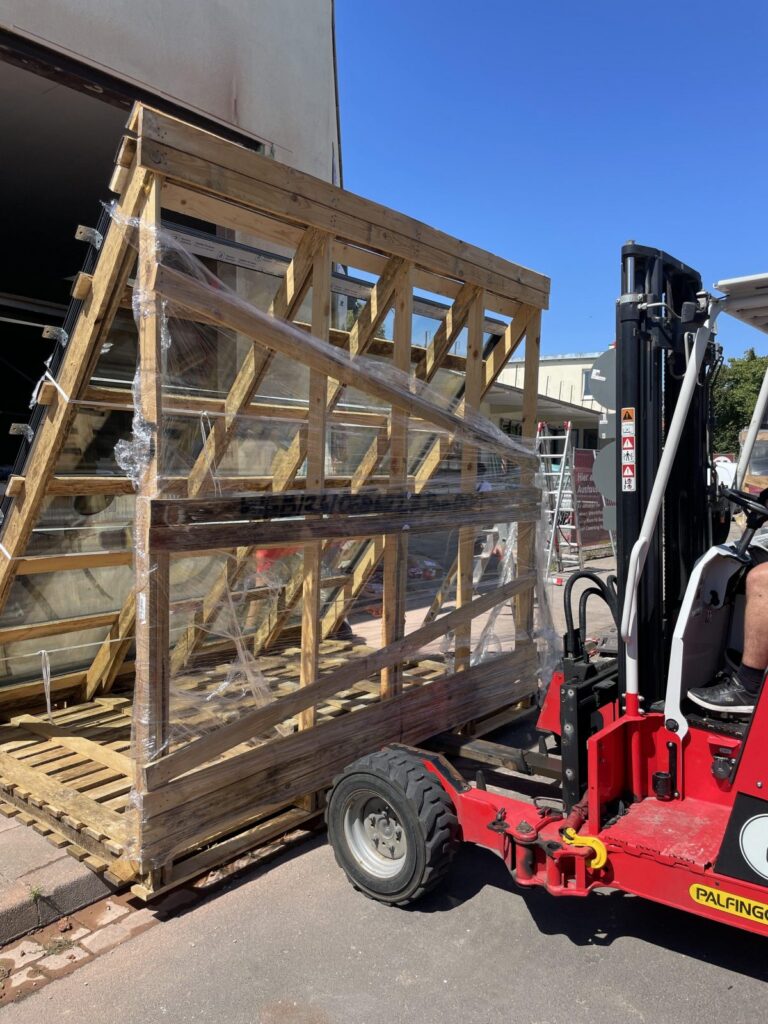 Ein Gabelstapler transportiert ein großes Fenster, das in einem Holzgestell mit Folie gesichert ist.