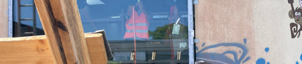 Ein großes Fenster wird in eine Fassadenöffnung eingesetzt. Hinter der Scheibe ist eine Person mit Warnweste zu sehen.
