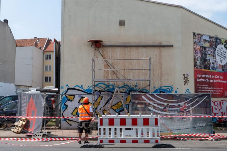 Eine Person mit Warnweste und Schutzhelm steht vor einer abgesperrten Hausfassade mit Gerüst und montierter Wandsäge.
