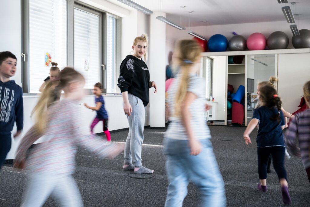 Kinder bewegen sich in einem Gymnastikraum, in der Mitte steht eine Trainerin.