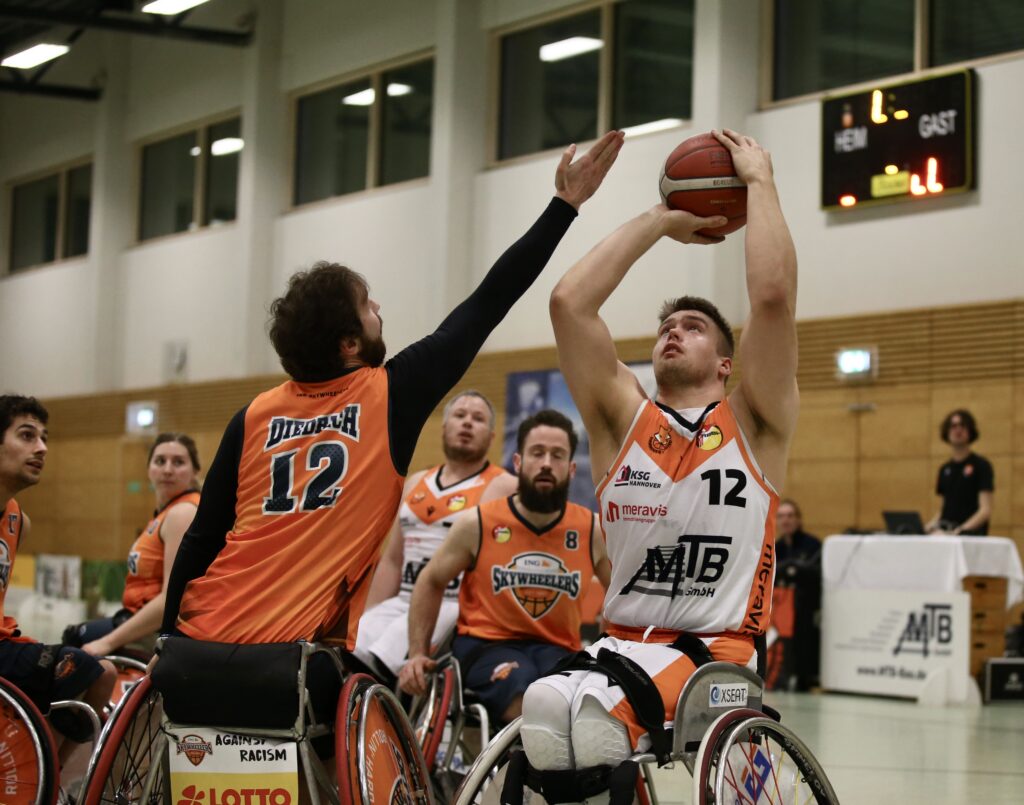 Zwei Rollstuhlbasketballer kämpfen in einer Sporthalle um den Ball, dahinter weitere Spieler.