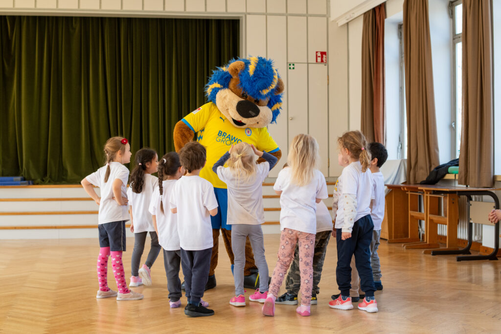 Kinder stehen in einer Turnhalle um ein Maskottchen in gelb-blauem Kostüm.