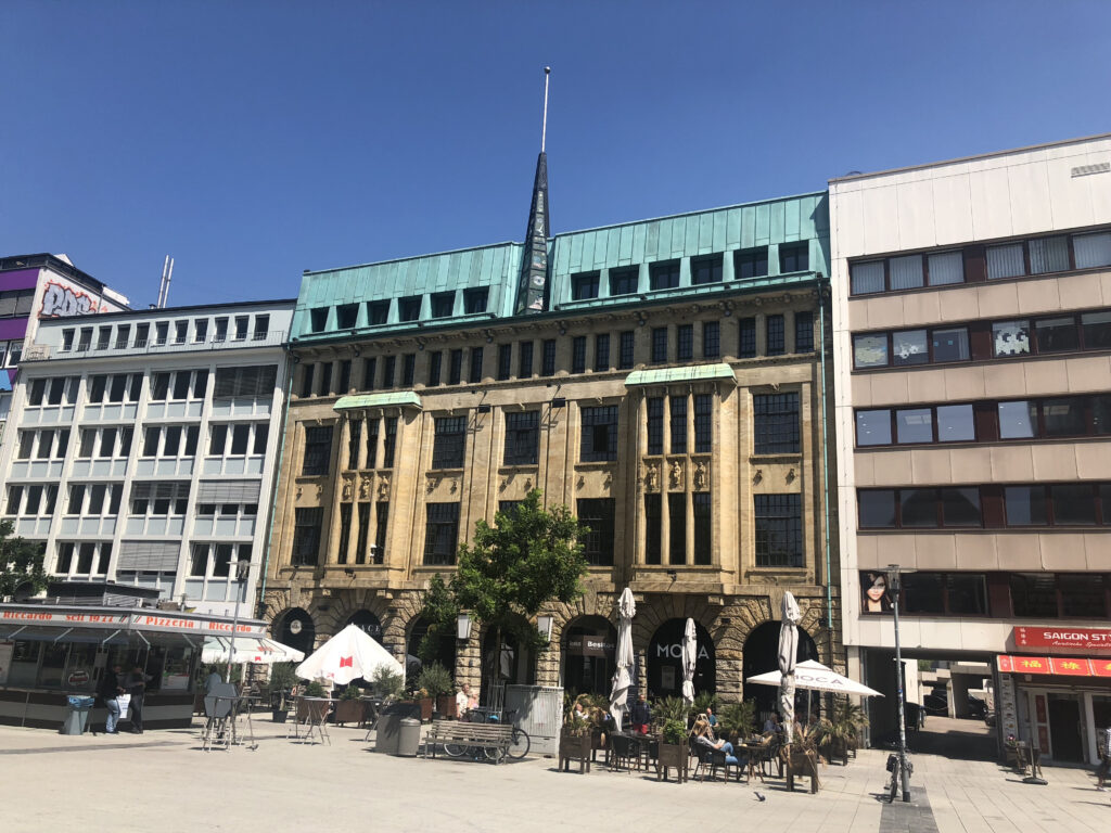 Außenansicht des Hauses mit grünem Kupferdach in Hannover, davor ein Platz mit Außengastronomie.