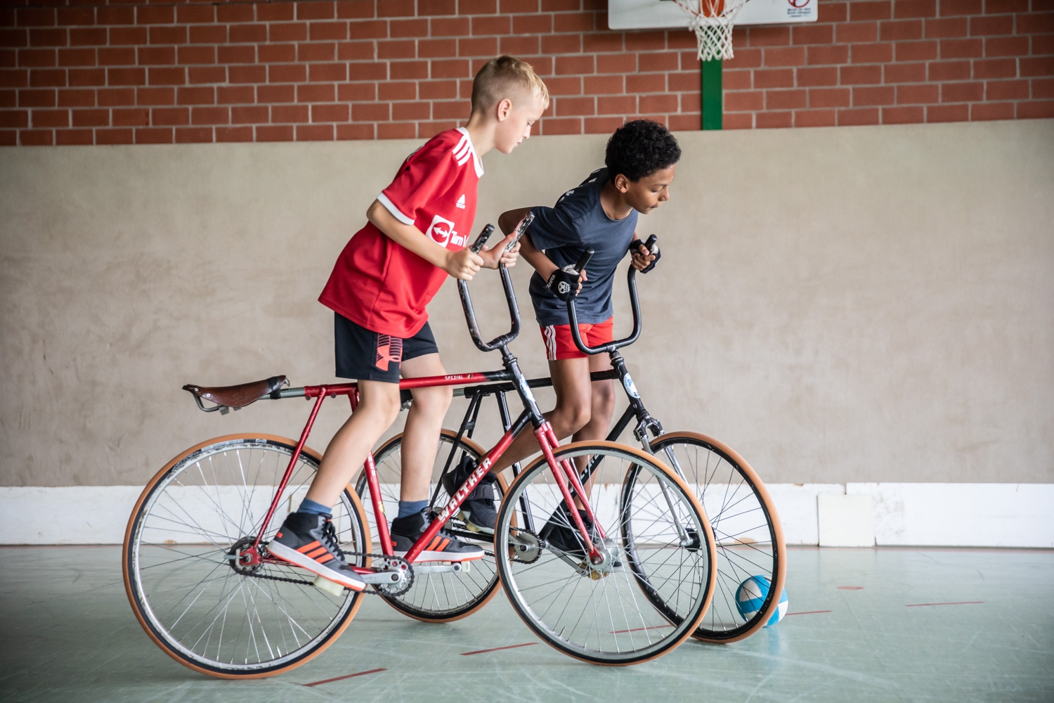 Zwei Jungen fahren in einer Sporthalle auf speziellen Fahrrädern einem Ball entgegen.