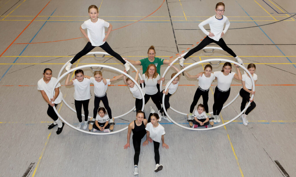 Gruppe von Mädchen in Sportkleidung formt mit weißen Röhnrädern eine symmetrische Figur in einer Sporthalle