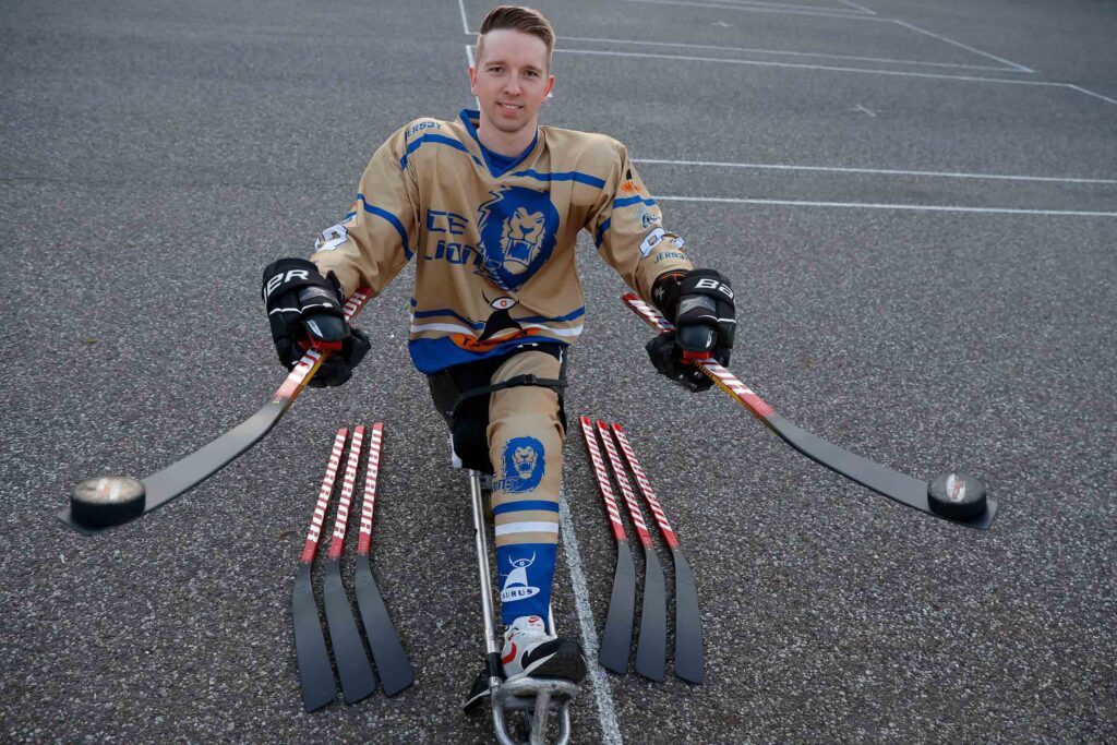 Ein sitzender Para-Eishockey-Spieler in kompletter Ausrüstung mit zwei Schlägern in den Händen und mehreren Schlägern vor sich auf dem Asphalt