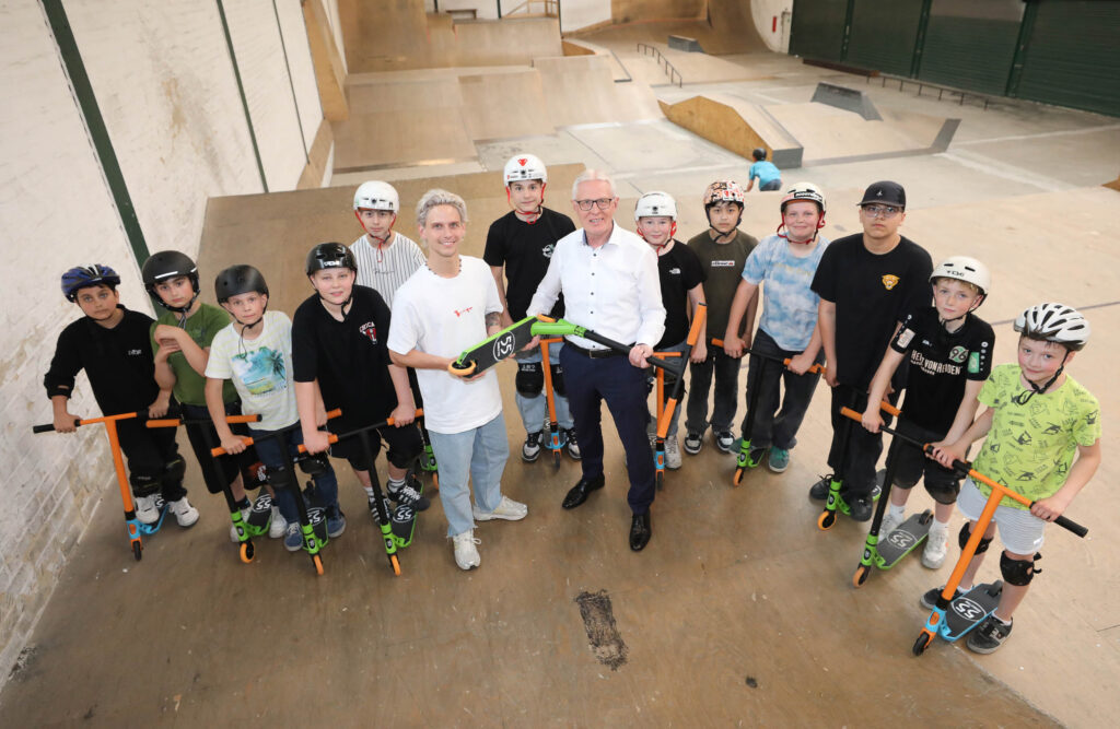 Gruppe von Kindern und zwei Erwachsenen mit Helmen und Scootern in einer Skatehalle.
