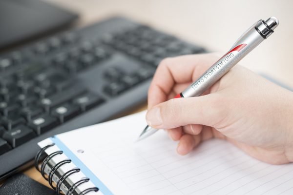 Eine Hand schreibt mit einem Stift in ein Notizbuch neben einer Tastatur.