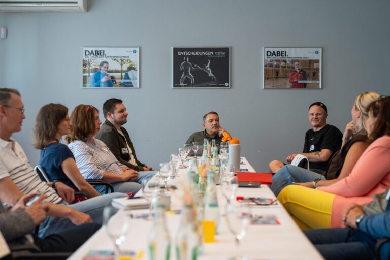 Mehrere Personen sitzen an einem langen Tisch im Gespräch. Auf dem Tisch liegen Unterlagen zwischen Gläsern und Getränkeflaschen.