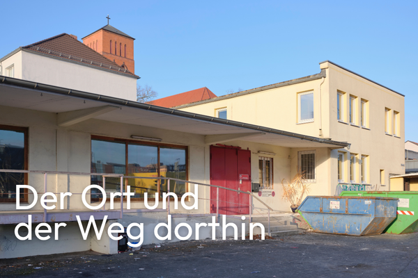 Ein flacher, hellgelber Gebäudekomplex mit Flachdach und mehreren Fenstern steht im Vordergrund. Links befindet sich eine Rampe mit Geländer, daneben eine große rote Tür. Rechts vor dem Gebäude steht ein blauer Container. Im Hintergrund ist ein weiteres Gebäude mit rotem Ziegeldach und einem kleinen Turm zu sehen. Der Himmel ist wolkenlos und blau. In der linken unteren Ecke steht weißer Text: 'Der Ort und der Weg dorthin'.