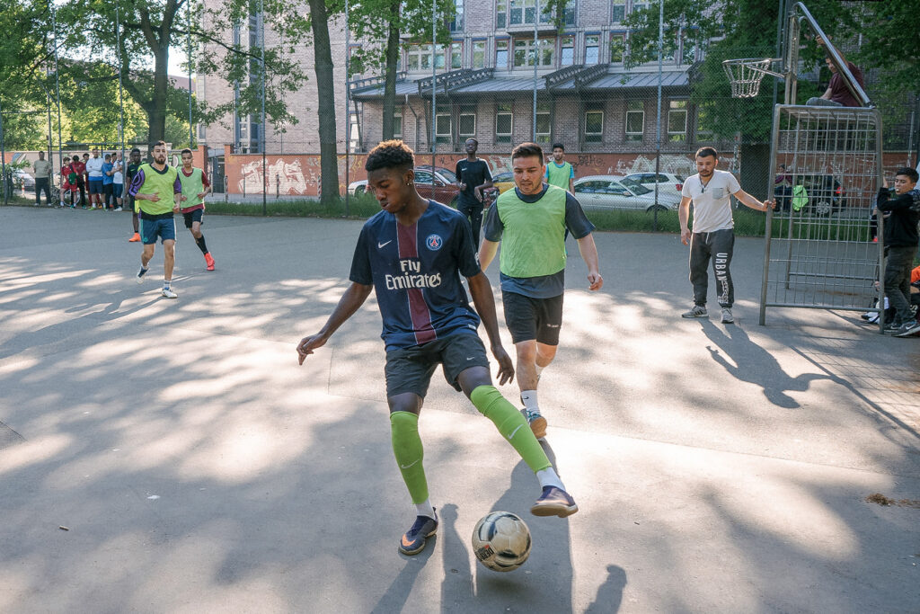 Junger Mann in dunklem Trikot spielt Fußball auf einem städtischen Platz, im Hintergrund Mitspieler.