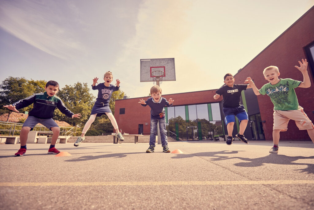 Kinder springen auf einem Schulhof, im Hintergrund ein Basketballkorb und ein Backsteingebäude.