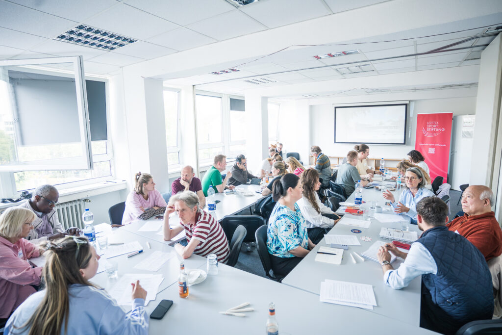 Menschen sitzen an zwei langen Tischen in einem hellen Besprechungsraum und führen eine Diskussion.