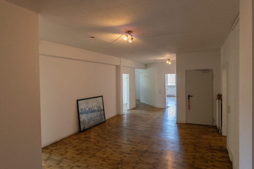Leerer Flur mit Holzboden, weißen Wänden, zwei Deckenlampen und einem großen Bildrahmen, der an der Wand lehnt.