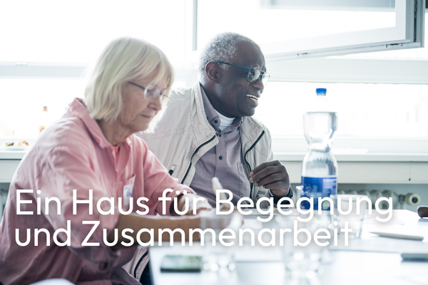 Zwei Personen sitzen im Gespräch an einem Tisch in einem hellen Raum. Unten steht der Text: „Ein Haus für Begegnung und Zusammenarbeit“.