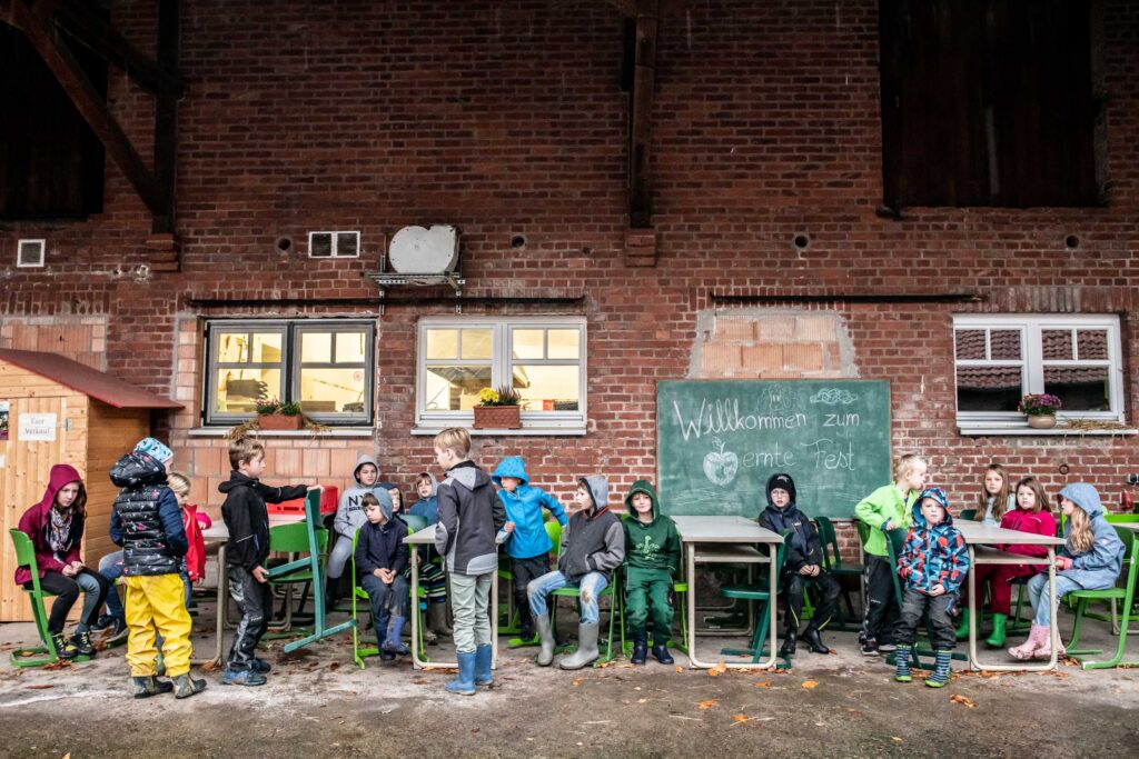 Eine Gruppe Kinder in wetterfester Kleidung versammelt sich vor einer Backsteinwand und einer Tafel mit der Aufschrift „Willkommen zum Erntefest“.