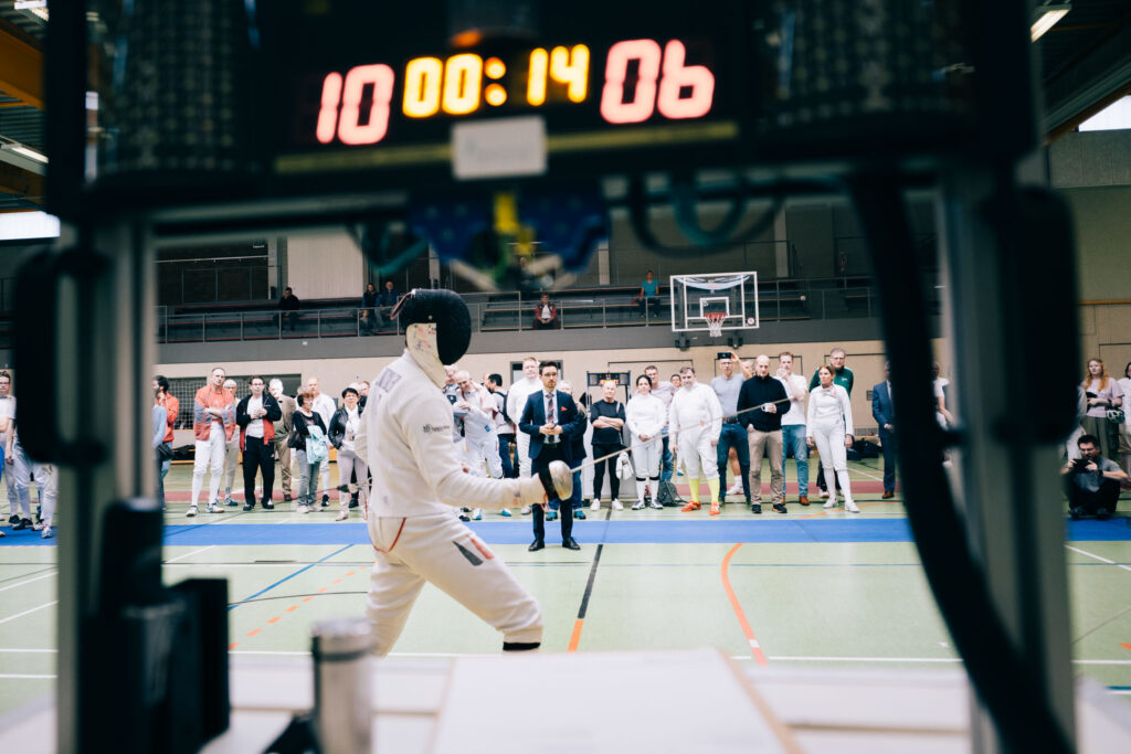 Blick durch eine digitale Anzeigetafel auf ein Fechtturnier in einer Sporthalle: Ein Fechter in weißer Schutzkleidung und Maske tritt vor Publikum und einem Schiedsrichter auf der Fechtbahn an.