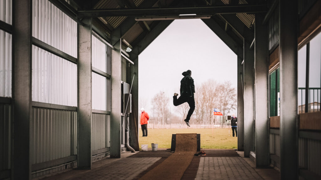 Person, die in einer überdachten Sportanlage einen dynamischen Sprung in der Luft beim Klootschiessen ausführt, im Hintergrund ist ein Sportplatz im Freien zu sehen.