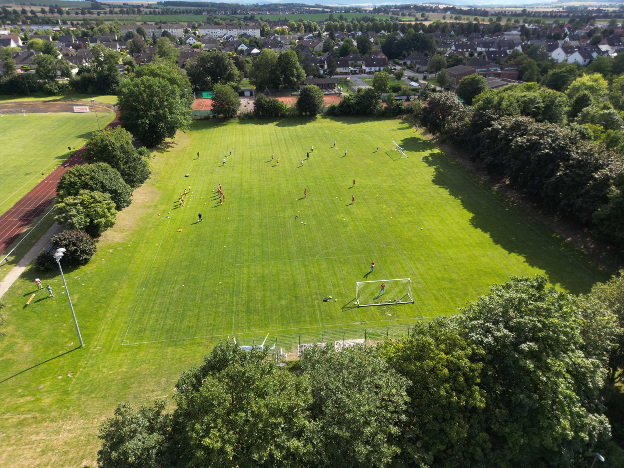 Vogelperspektive auf einen großen grünen Rasen-Sportplatz mit verteilt trainierenden Personen, Toren und einer angrenzenden Laufbahn am Rande eines Wohngebiets.