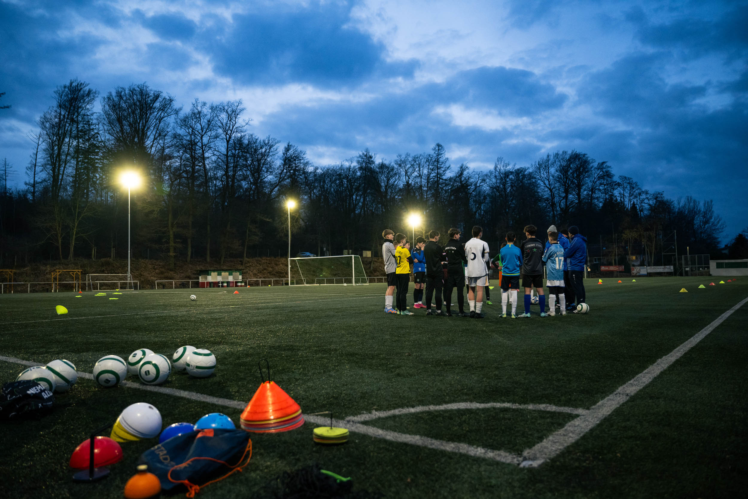 Eine Jugend-Fußballmannschaft versammelt sich bei Dämmerung unter Flutlicht zu einer Besprechung auf dem Kunstrasenplatz, während im Vordergrund Trainingsutensilien wie Fußbälle und Markierungshütchen liegen.