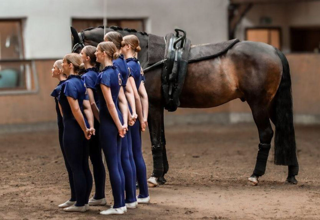 Eine Gruppe von sechs Voltigiererinnen in blauen Trikots steht aufgestellt neben einem Pferd in einer Reithalle.