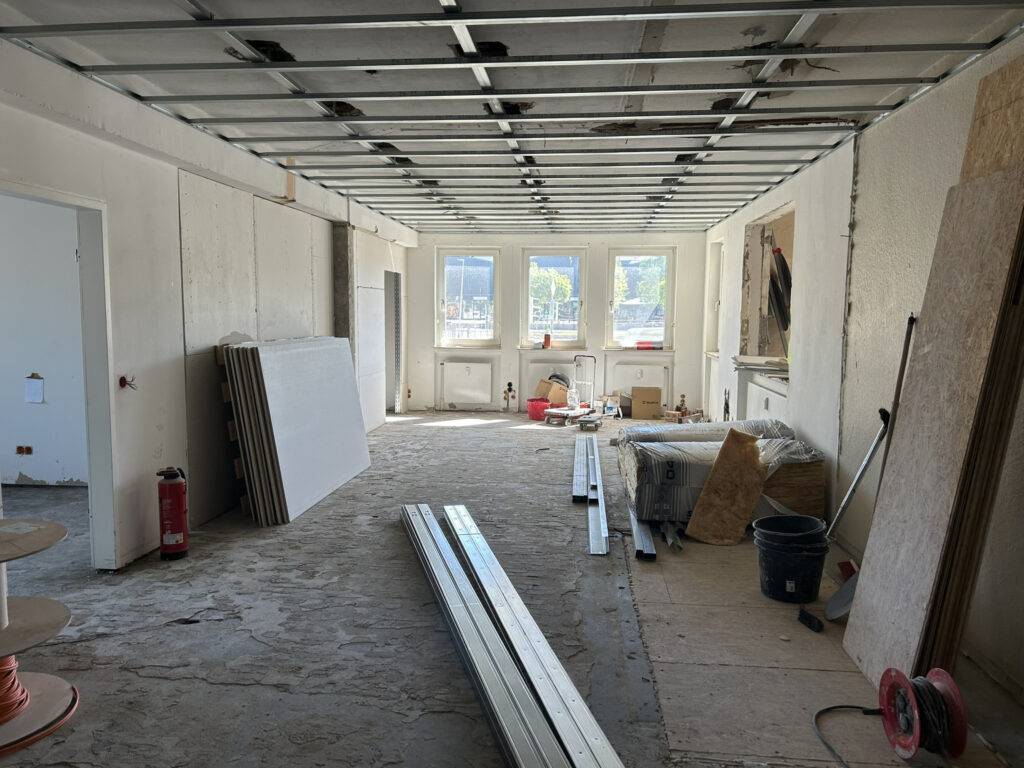 Raum im Umbau mit freiliegendem Deckengitter, angelehnten Gipskartonplatten, Metallprofilen am Boden und Fenstern am Ende.